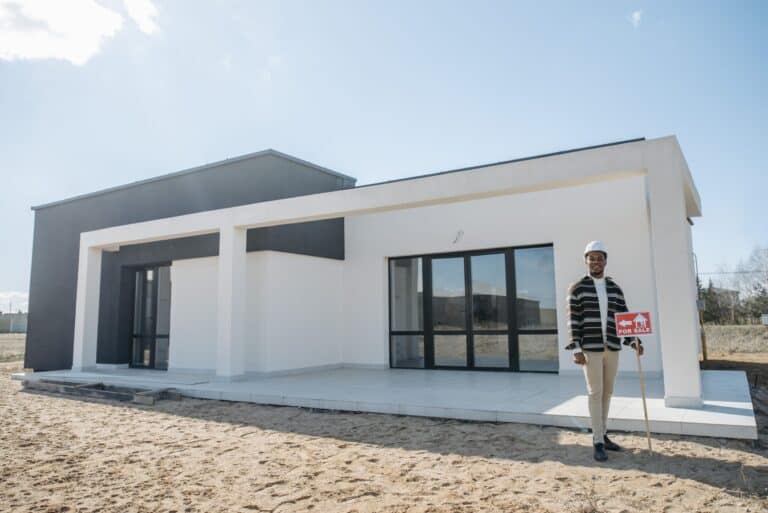 pexels-photo-7937765 - Stonebrook Custom Homes Man in a hard hat stands beside a modern white house under construction, holding a red 'For Sale' sign on a sunny day. | Stonebrook Custom Homes