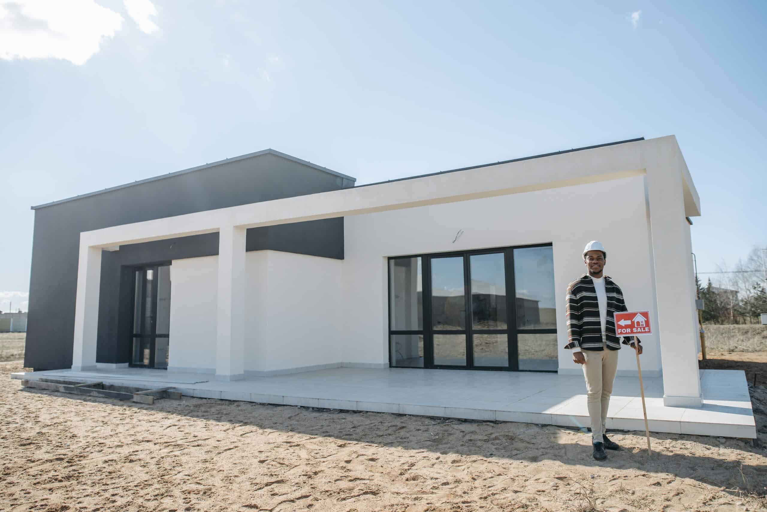 pexels-photo-7937765 - Stonebrook Custom Homes - Stonebrook Custom Homes Man in a hard hat stands beside a modern white house under construction, holding a red 'For Sale' sign on a sunny day. | Stonebrook Custom Homes | Stonebrook Custom Homes