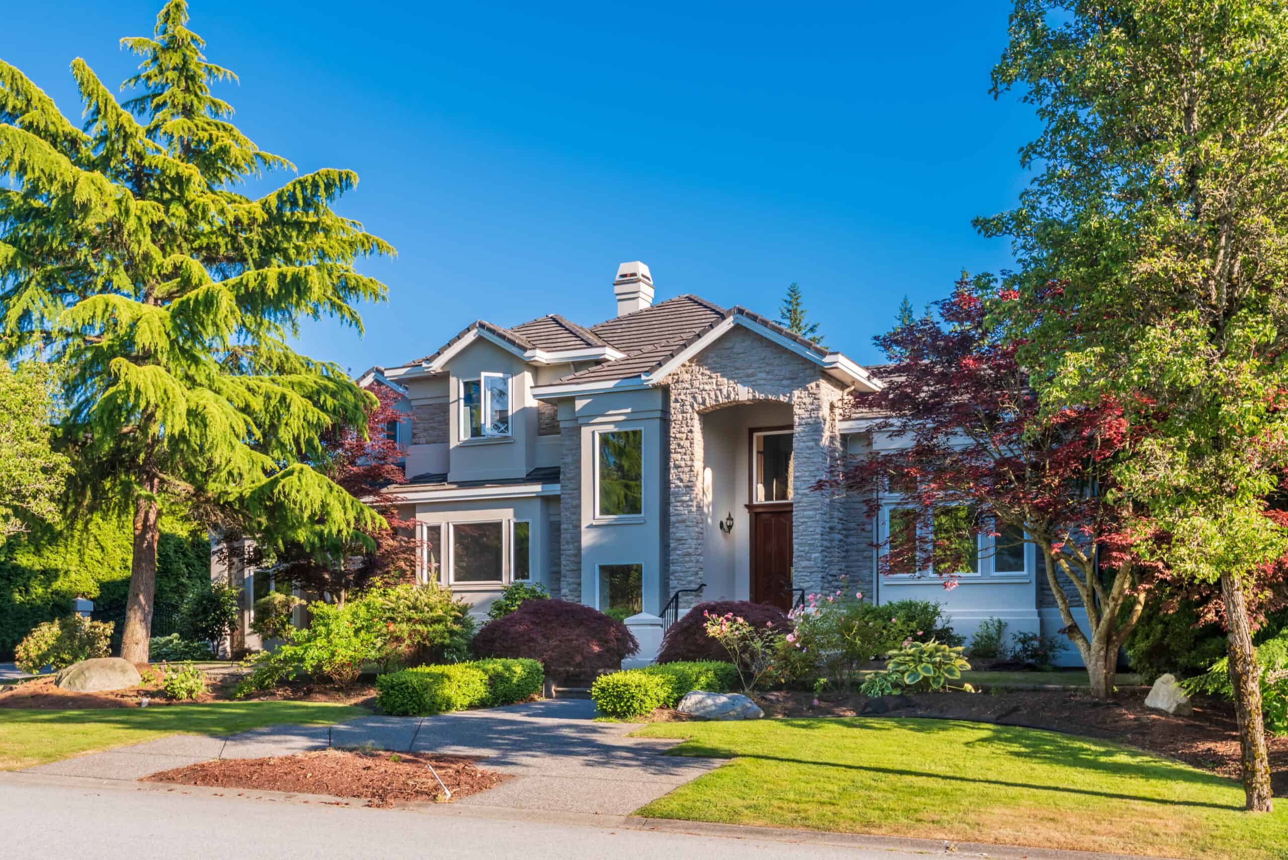 beautiful-exterior-of-newly-built-luxury-home-yar-2025-10-28-21-41-54-utc - Stonebrook Custom Homes - Stonebrook Custom Homes Two-story house with a stone facade and arched entry, surrounded by lush landscaping and tall trees under a clear blue sky. | Stonebrook Custom Homes | Stonebrook Custom Homes